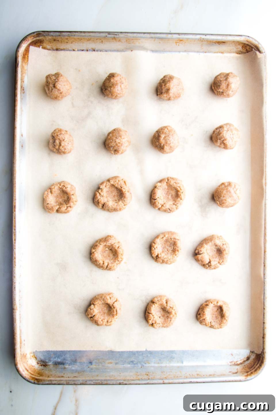 Large rimmed baking sheet, lined with parchment paper, filled with rows of cookie dough rolled into balls. Some of the cookies have a thumbprint indent.
