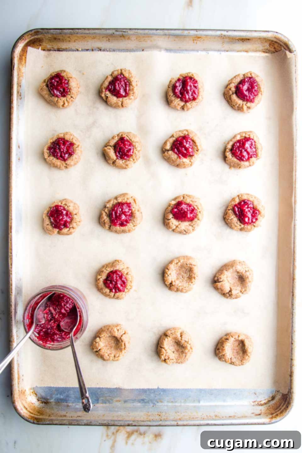 Large rimmed baking sheet, lined with unbleached parchment paper, lined with neat rows of thumbprint cookies. Half are filled with cranberry jam, there is a small glass dish of jam with two spoons in it, for filling the remaining cookies.