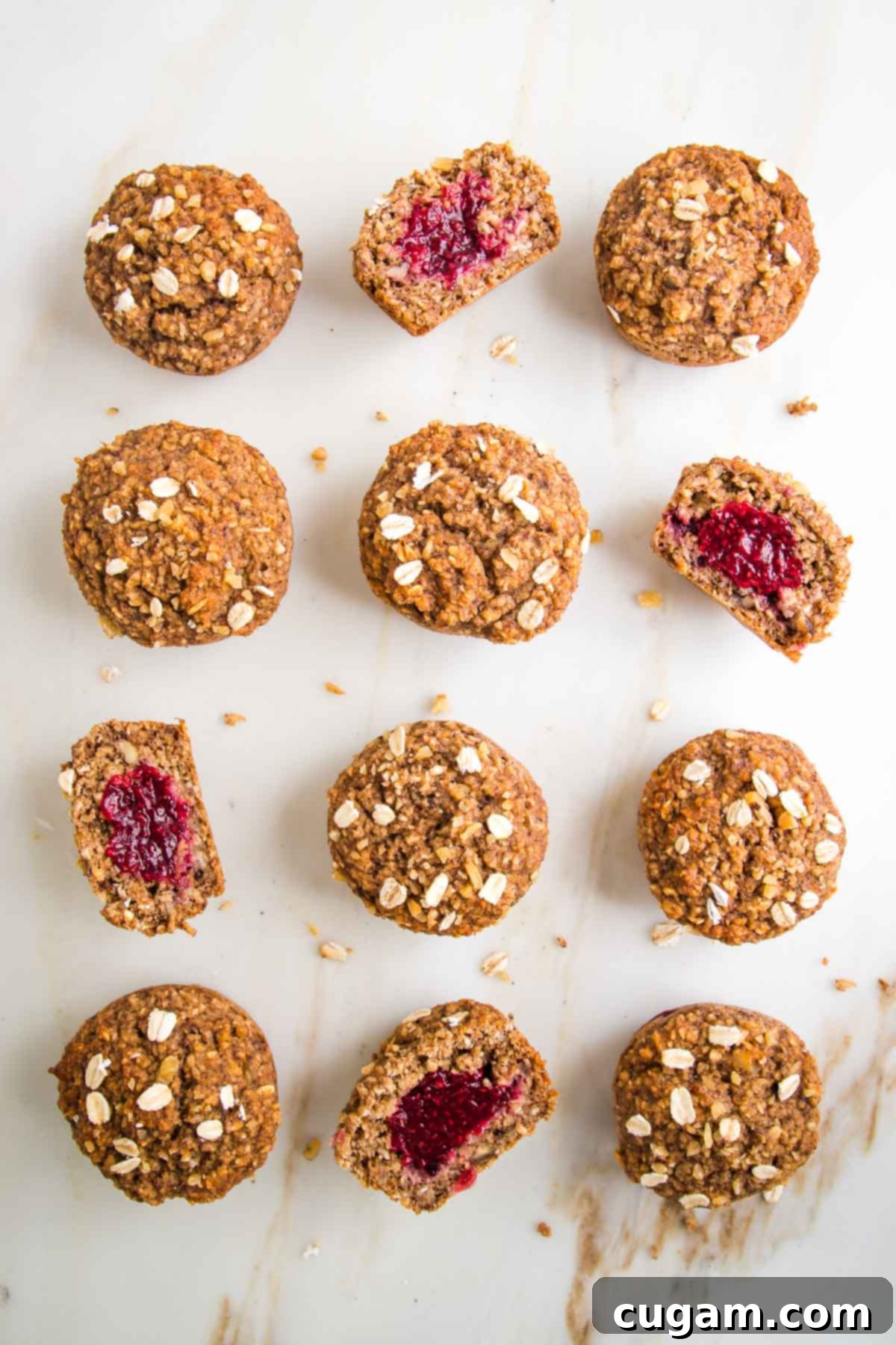 A close-up of a dozen healthy oatmeal muffins, each generously filled with jam. Several muffins are cut in half to showcase the delightful jam filling within.