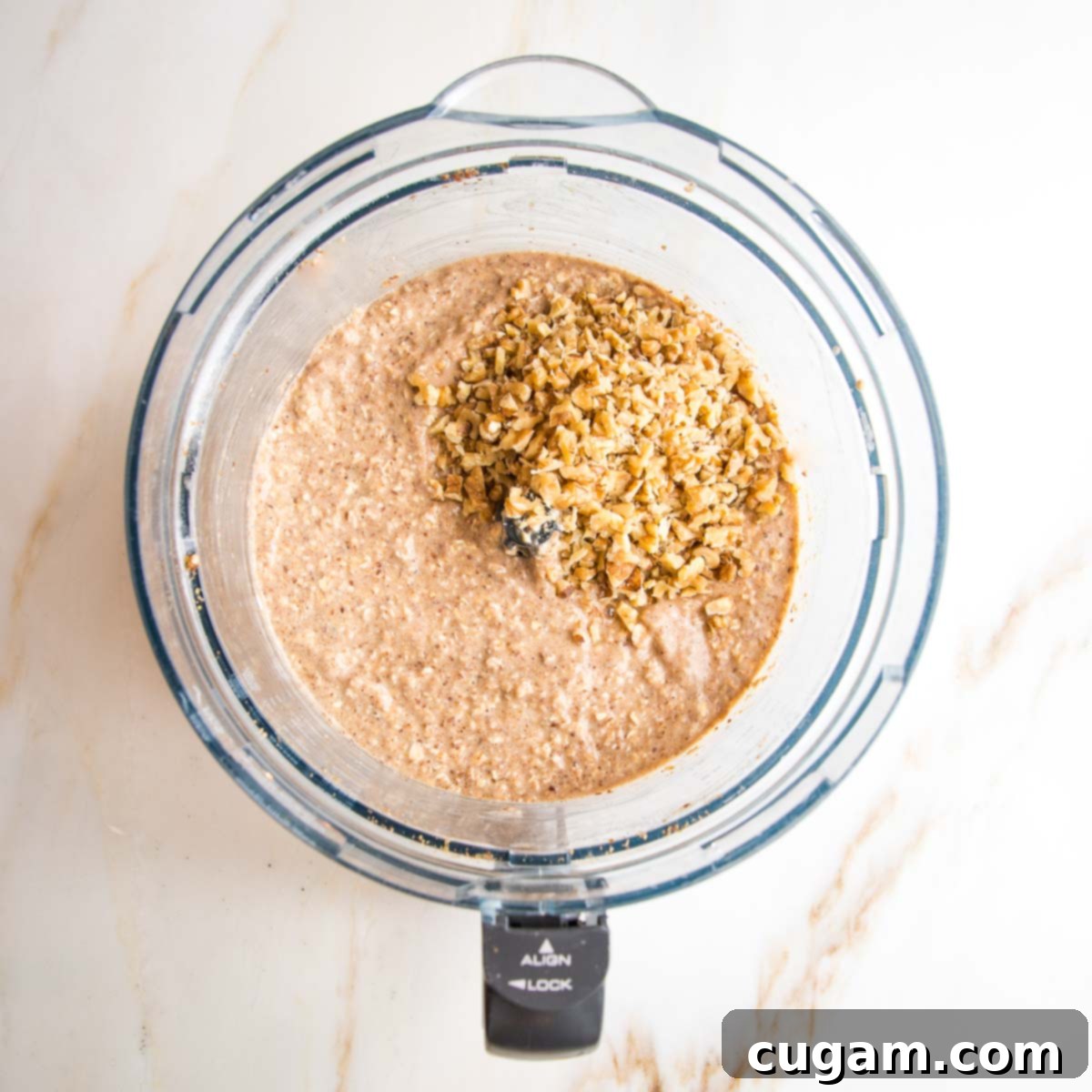Smooth oatmeal muffin batter in a food processor bowl, topped with a generous scoop of finely chopped walnuts, ready to be folded in.