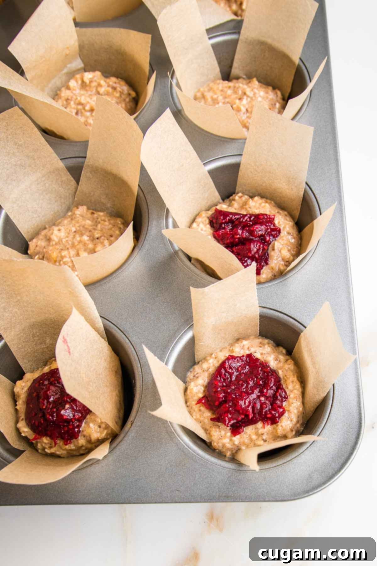 A muffin tin with cups half-filled with oatmeal batter, each topped with a dollop of red jam, ready to be covered with the remaining batter.