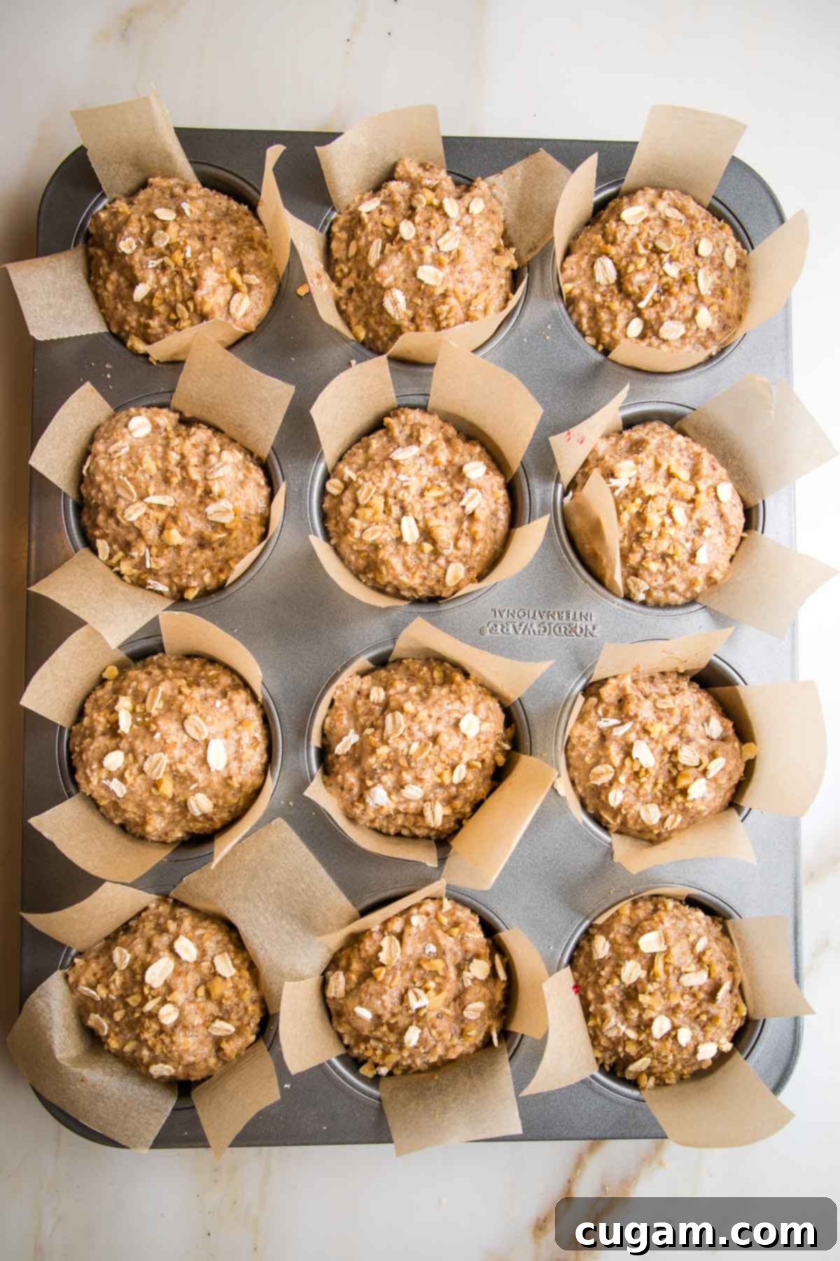 A muffin tin, lined with parchment strips, with each cup filled to the brim with oatmeal batter, covering the jam filling, ready for baking.