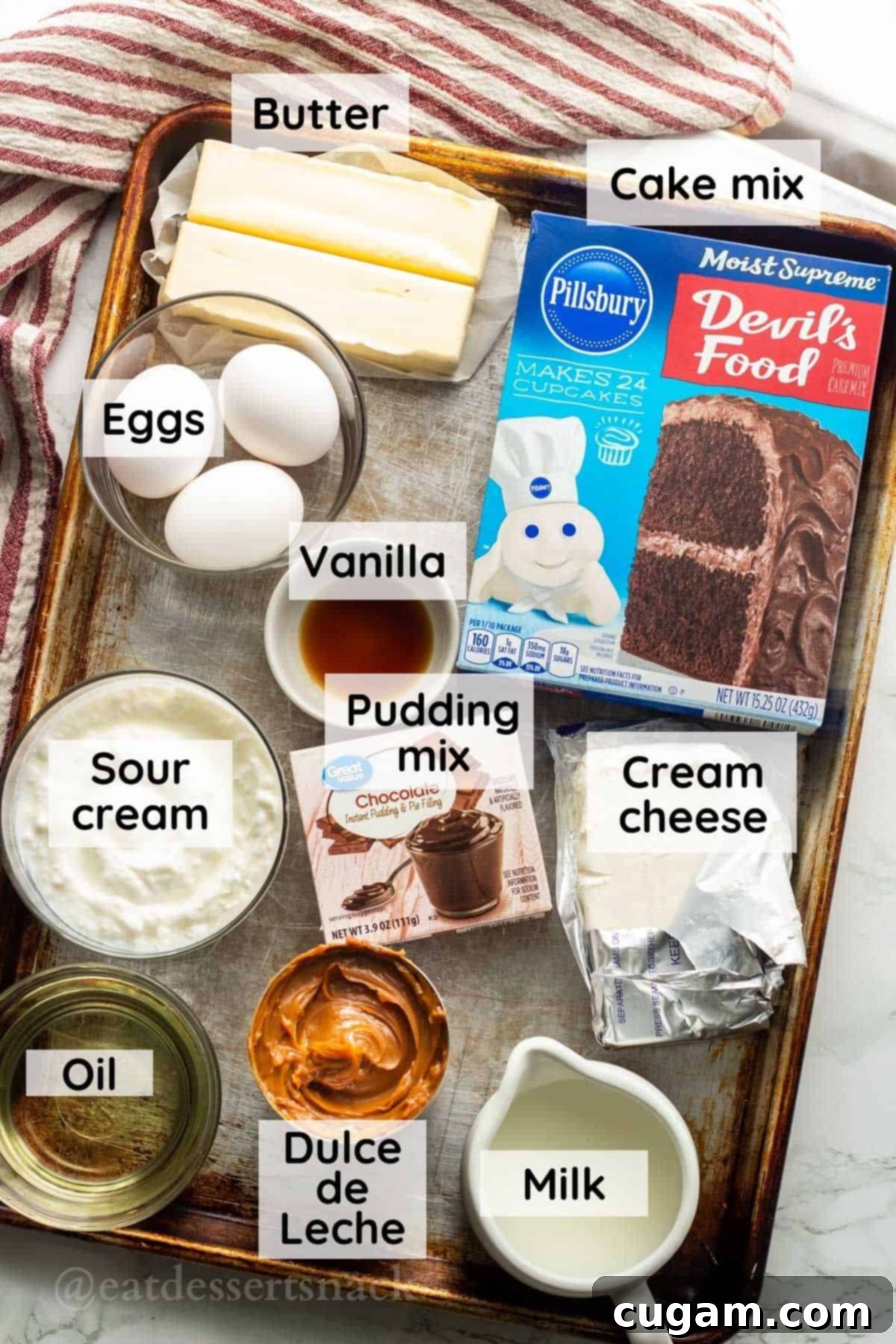 Decadent Chocolate Caramel Cupcakes 3 Selection of key ingredients including butter, eggs, vanilla, chocolate pudding mix, cake mix, cream cheese, milk, oil, and caramel, laid out on a metal baking sheet.