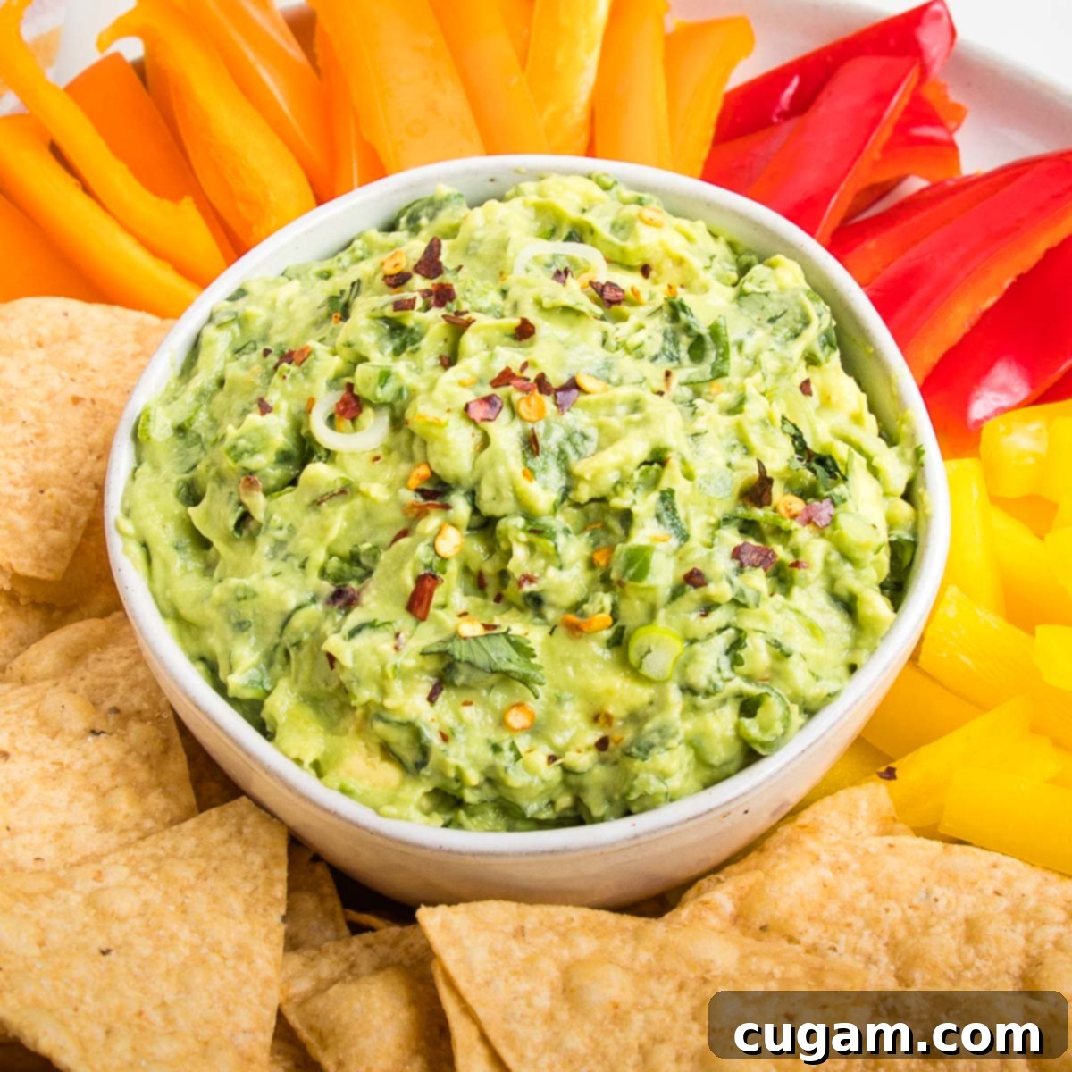 Close up of a bowl of chunky vegan guacamole sprinkled with red pepper flakes. The bowl is surrounded by tortilla chips and assorted color strips of bell peppers, perfect for game day dipping.