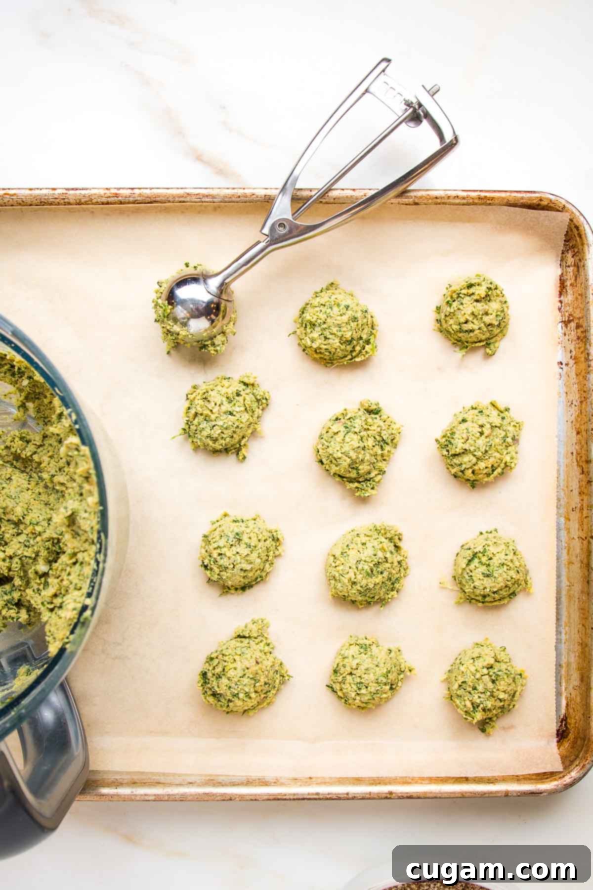 Large rimmed baking sheet, lined with unbleached parchment paper. You can see the far edge of a food processor bowl with a greenish pasty falafel mixture.  A small scoop is making golf ball sized portions onto the tray.