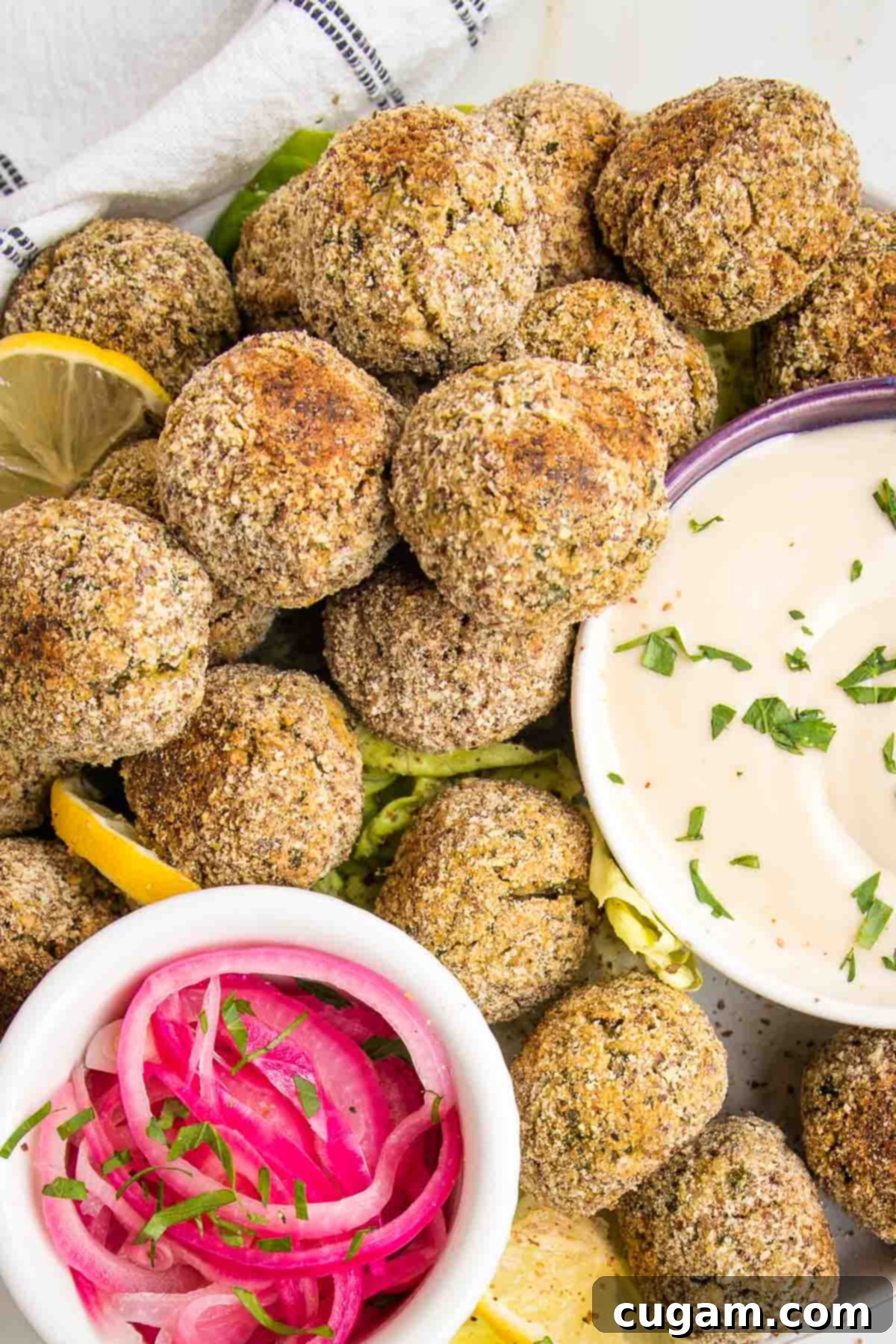 Plate filled with falafel balls and a small white dish with red pickled onions and another dish with tahini sauce sprinkled with fresh green herbs.