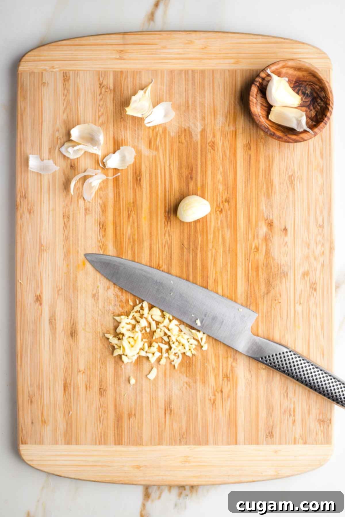 Cutting board, showing garlic cloves in various stages of peeled, chopped, in a bowl.