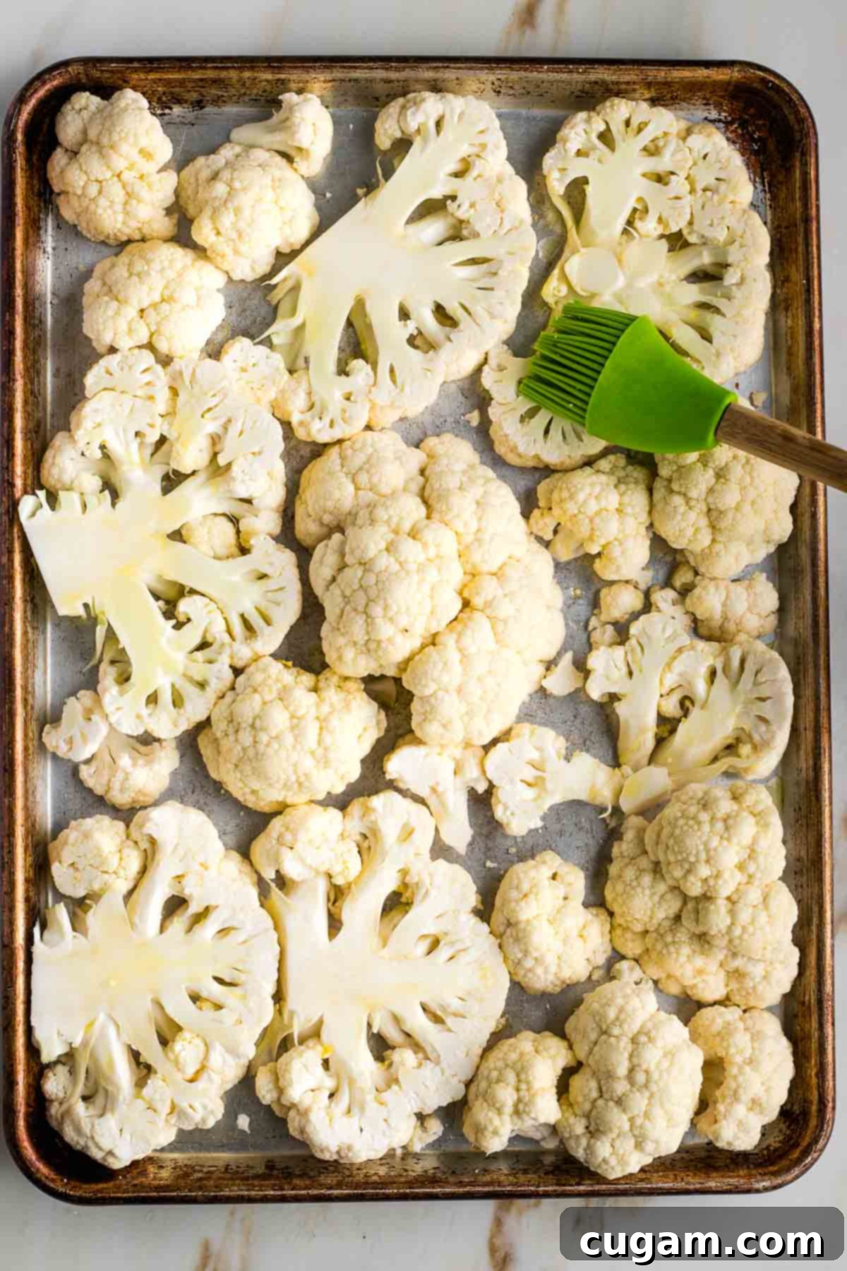 A pastry brush applying a thin layer of olive oil onto cauliflower steaks on a rimmed baking tray, ensuring even coating for crispiness.