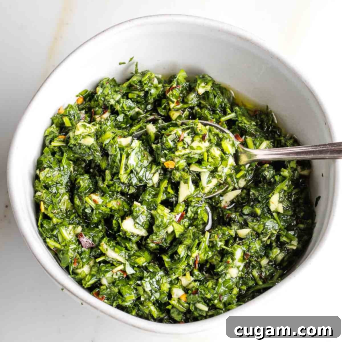 Bright green chimichurri sauce, vibrant with garlic and crushed red pepper, in a small bowl with a spoon, ready to be served.