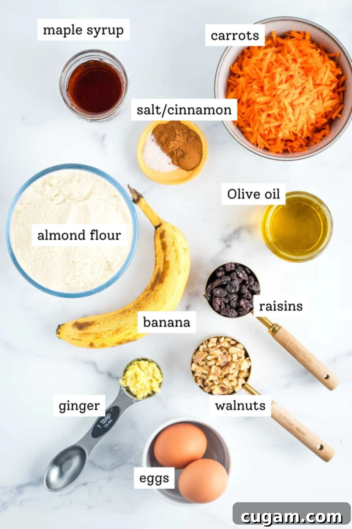 A selection of fresh ingredients including bananas, eggs, olive oil, maple syrup, grated carrots, ginger, raisins, walnuts, cinnamon, salt, and almond flour, laid out for baking.