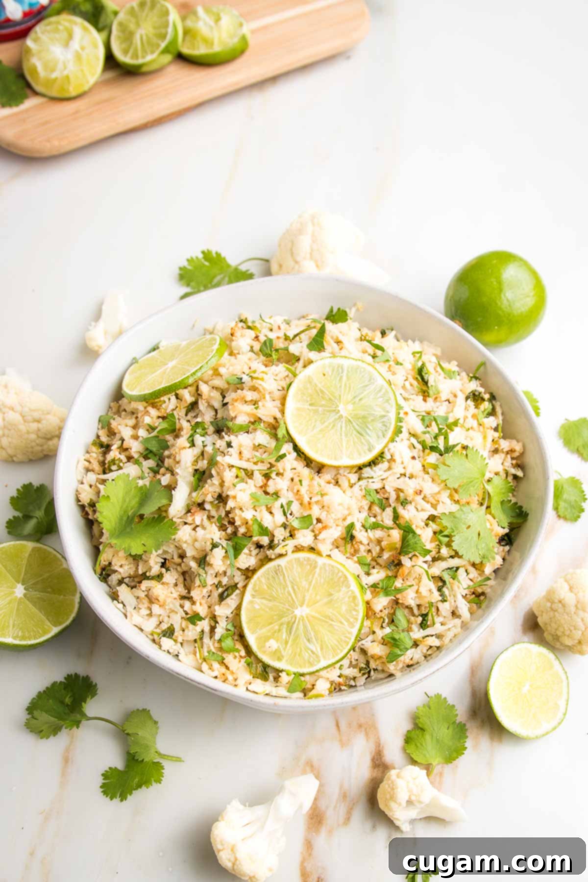 Effortless Stovetop Cauliflower Rice 2 Cauliflower rice in a white bowl with limes and fresh cilantro. A sprinkling of ingredients around the bowls edge.
