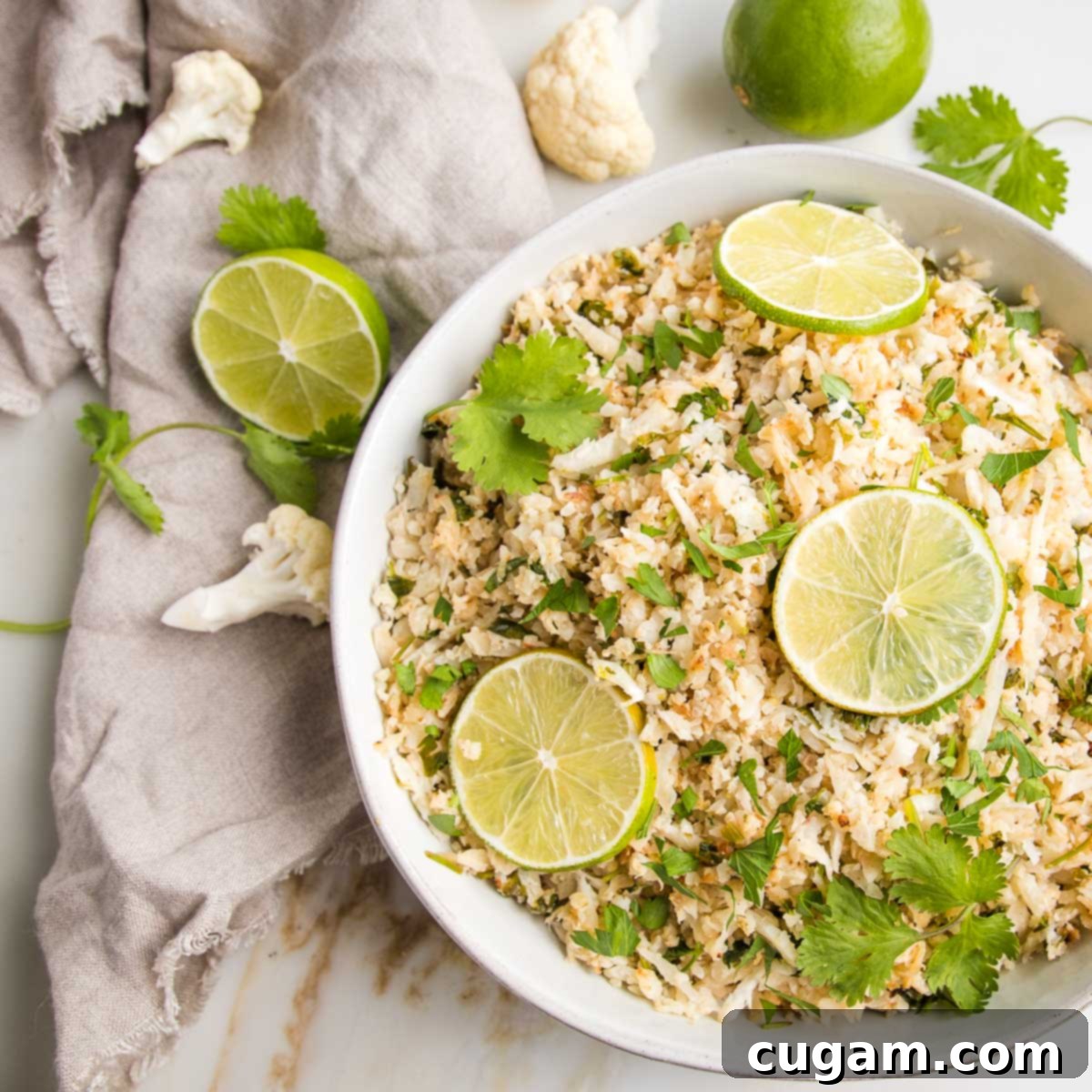 Effortless Stovetop Cauliflower Rice 12 White bowl filled with cauliflower rice, topped with fresh cilantro sprigs and slices of limes. A beige linnen napkin is on the side with slices of lime, florets of cauliflower and fresh cilantro.