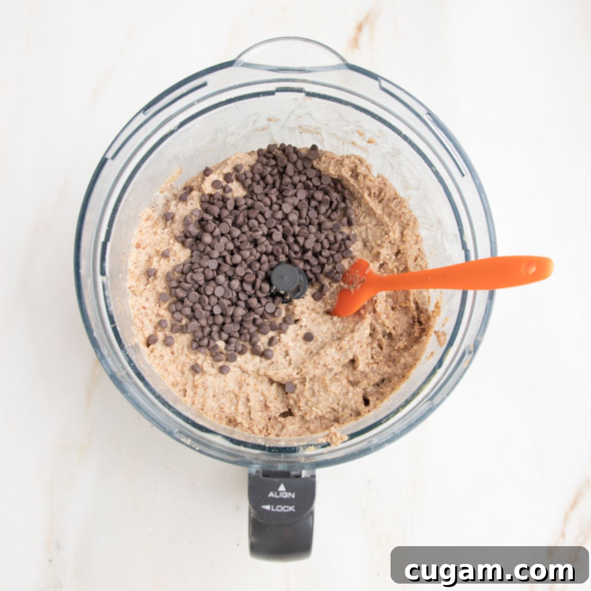 Banana bread batter in bowl of food processor with an orange spatula mixing in chocolate chips by hand.