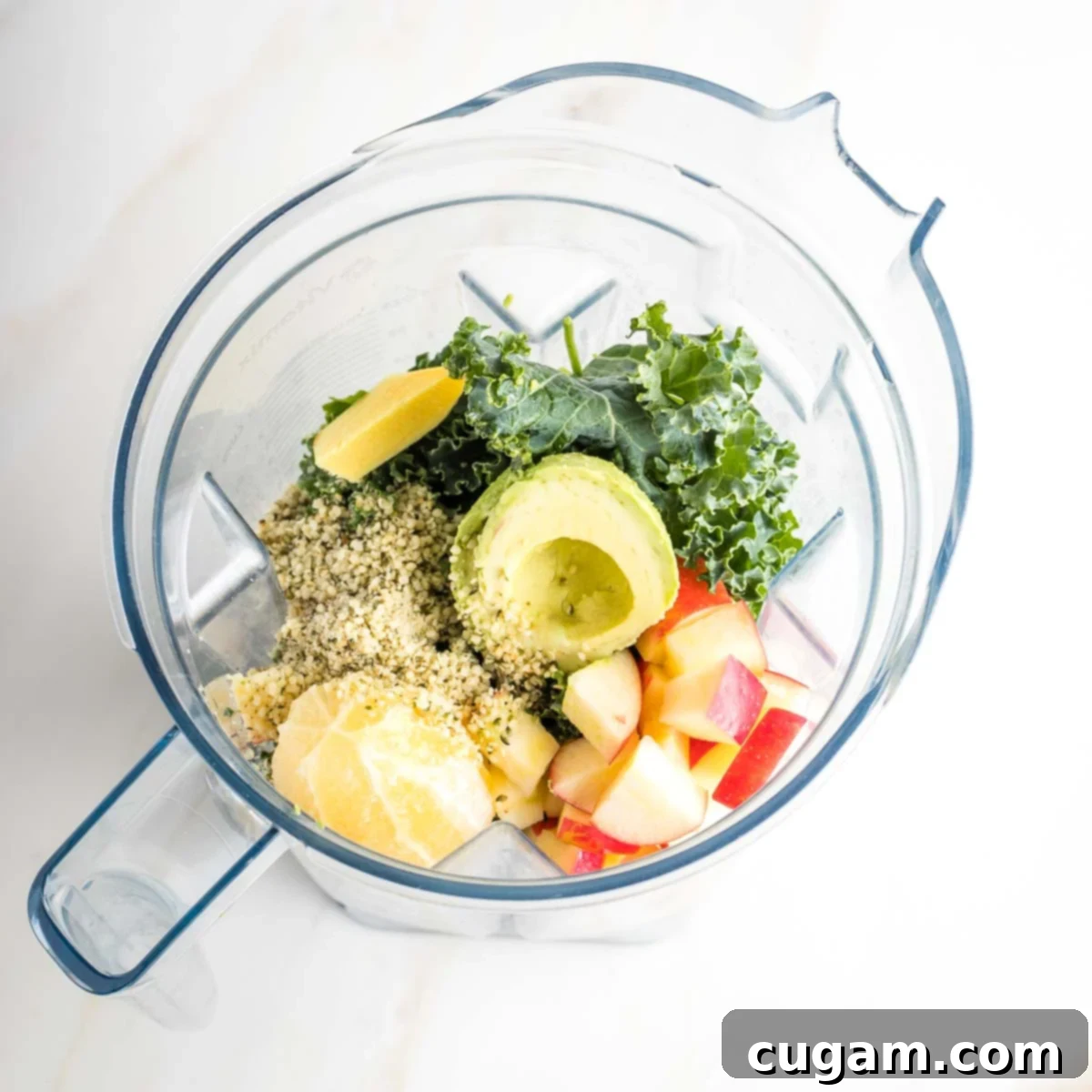Avocado half, kale, spinach, lemon, ginger, apple and hemp seeds in the pitcher of a blender.