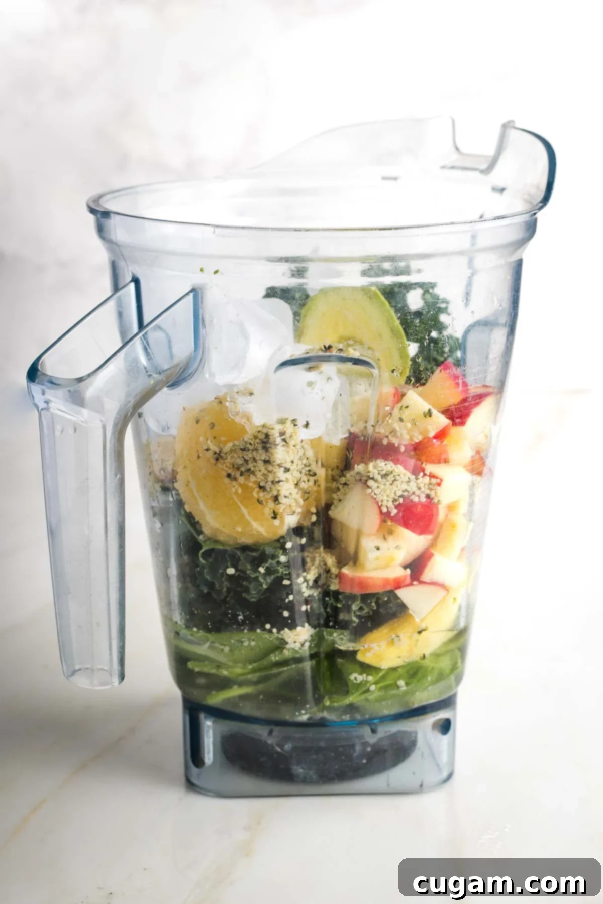 Blender pitcher filled with avocado kale smoothie ingredients including ice, hemp seeds, lemon.
