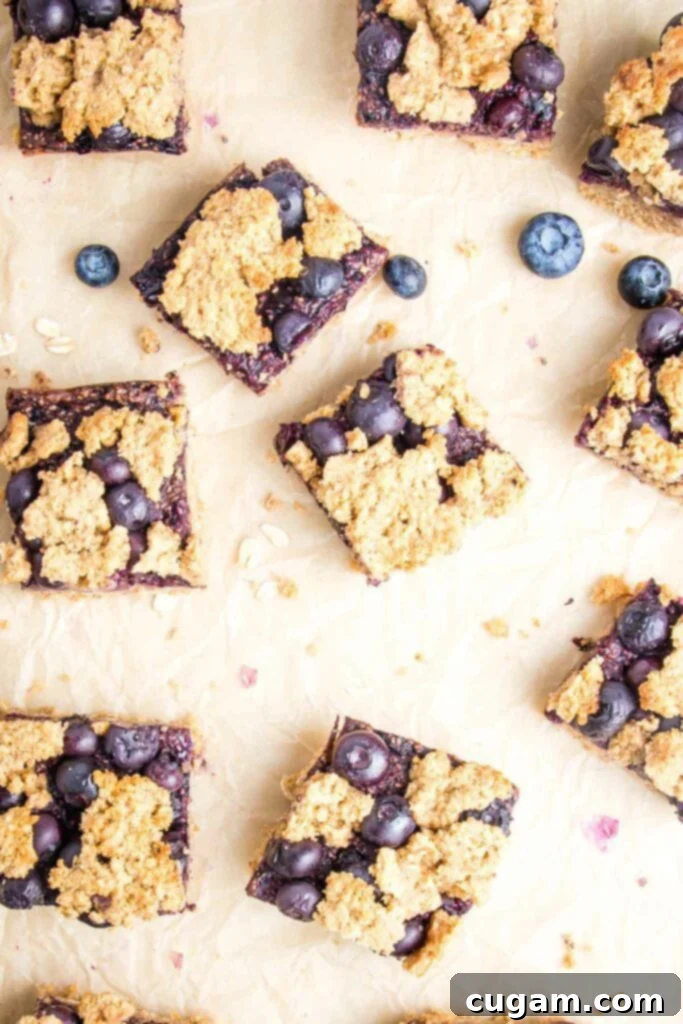 Blueberry Crumb Bars cut into squares on a piece of parchment paper with a few fresh blueberries sprinkled about. Showing a golden, crispy topping and a hint of the jammy filling.