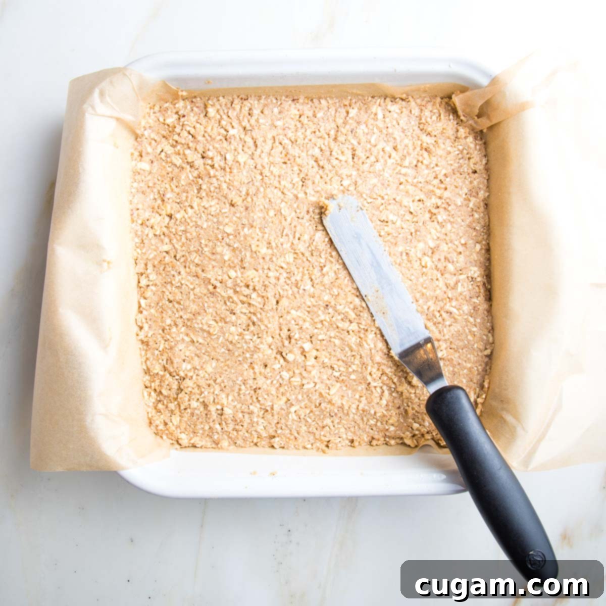 Offset spatula spreading batter into the corners of a square baking pan, creating an even base.