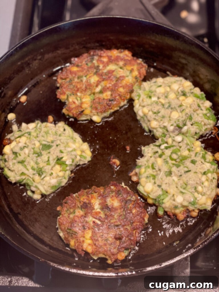 Large cast iron skillet with 5 corn and zucchini fritters frying. 2 have already been flipped and show golden brown, the other 3 shows the shredded zucchini batter.