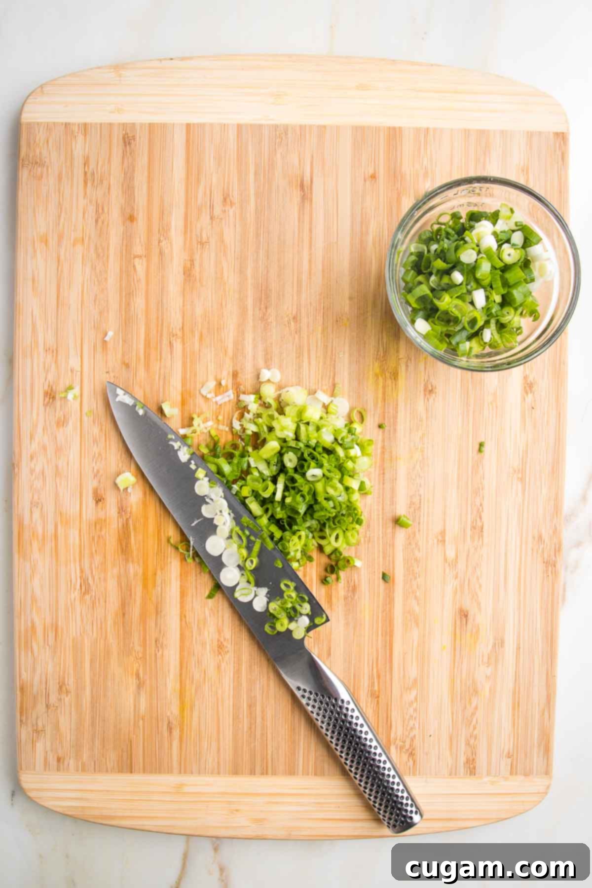 Wooden cutting board and sharp knife thinly slicing scallions with a small bowl off to the side with already sliced green onions.