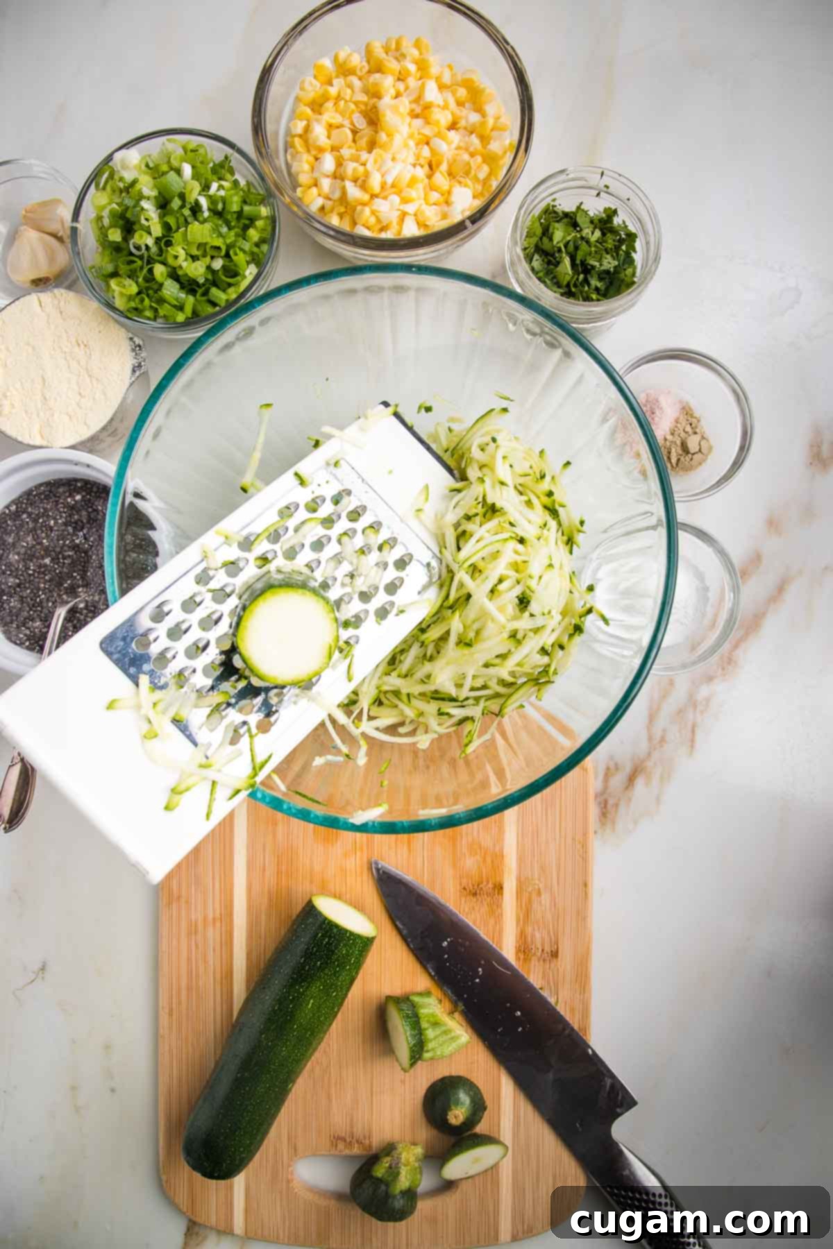Large mixing bowl with side of box grater, showing shredded zucchini into the bowl. Other ingredients for vegetable fritters shown around the bowl.