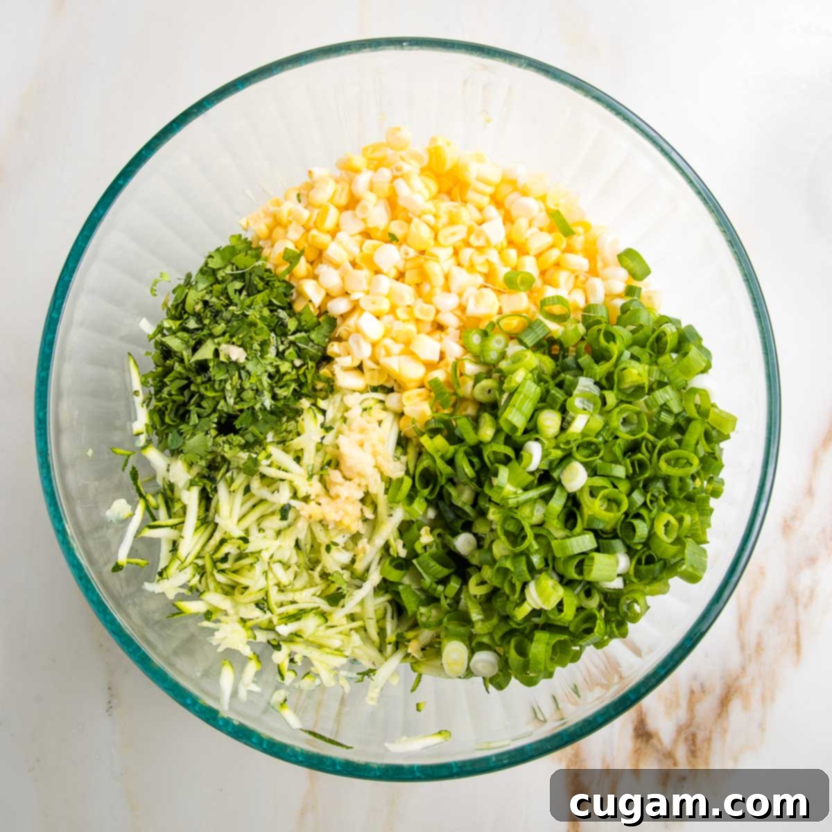 Ingredients for fritters in a glass bowl: corn kernels, grated zucchini, sliced scallions, chopped cilantro and grated garlic.