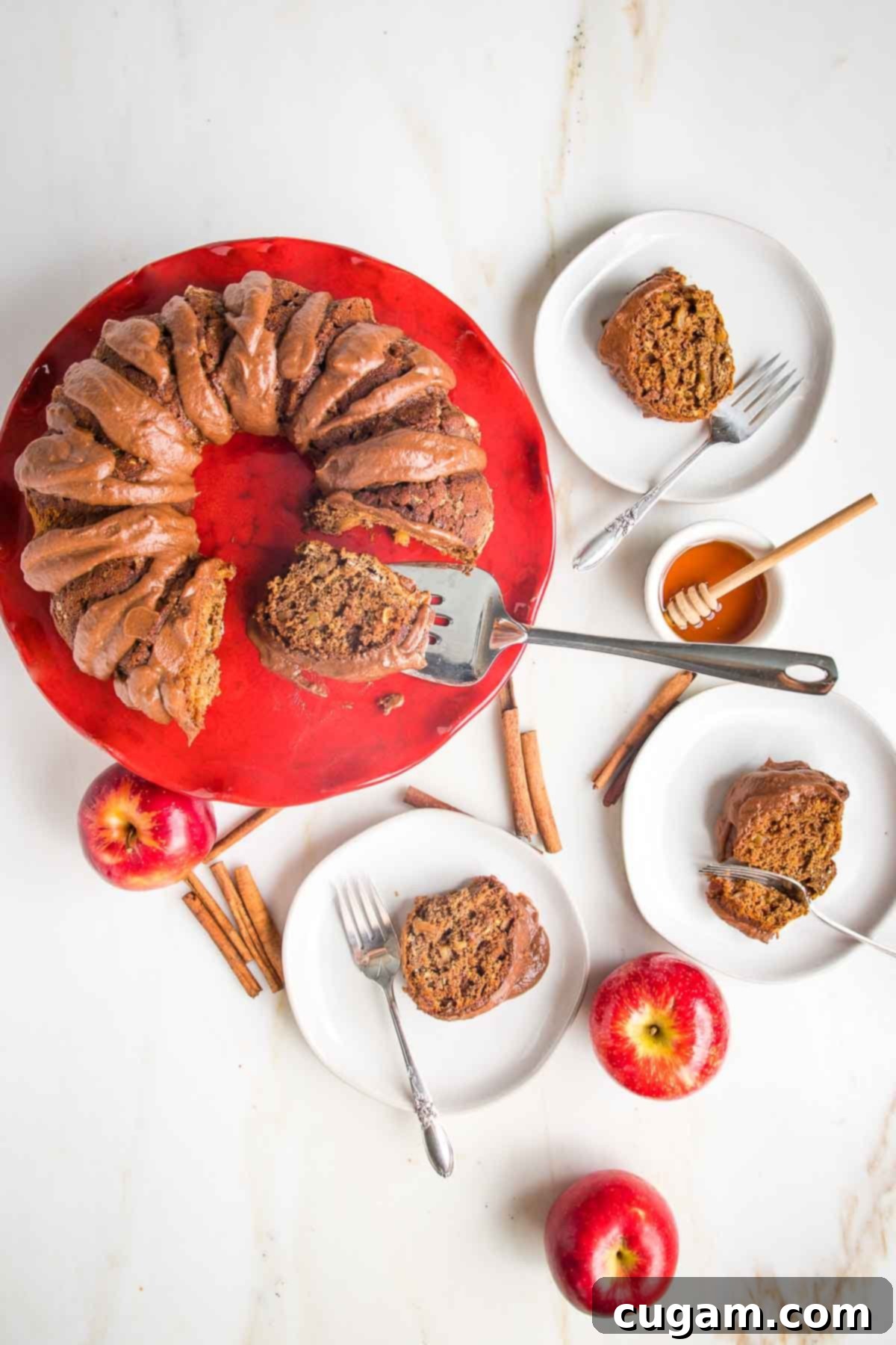 A vibrant red pedestal holding a golden apple cinnamon bundt cake, elegantly drizzled with chocolate sauce. Surrounding the cake are small plates with individual cake slices, a small dish of golden honey, and fragrant cinnamon sticks, all set against a warm, inviting counter. This scene perfectly captures the cozy essence of fall baking and holiday celebrations.