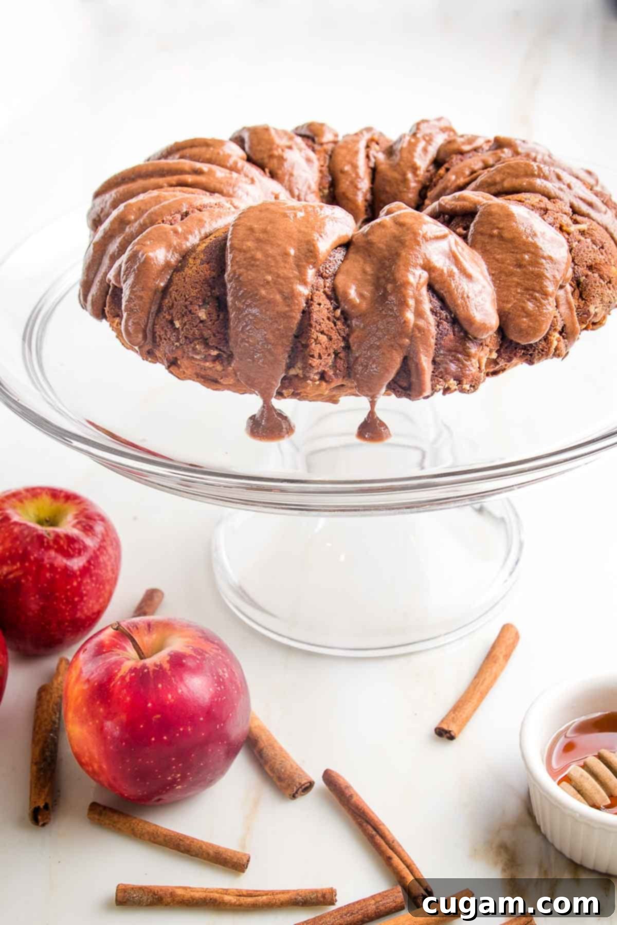 A magnificent apple bundt cake, glistening with a rich chocolate drizzle that elegantly drips down its sides, proudly displayed on a sleek glass pedestal. Surrounding the pedestal are whole, fresh red apples, fragrant cinnamon sticks, and a small, inviting bowl of golden honey, all contributing to a warm, autumnal ambiance.