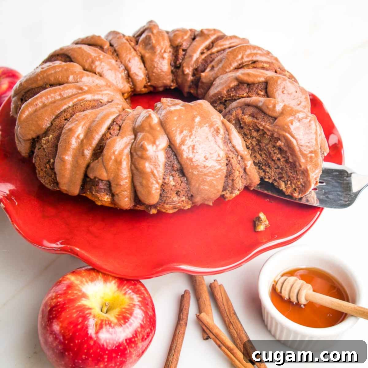 A beautifully presented apple bundt cake, generously coated with a dark chocolate drizzle, takes center stage on a striking red cake stand. It is artfully surrounded by a whole, ripe apple, fragrant cinnamon sticks, and a small, inviting bowl of golden honey, creating a warm and appealing autumn harvest scene.