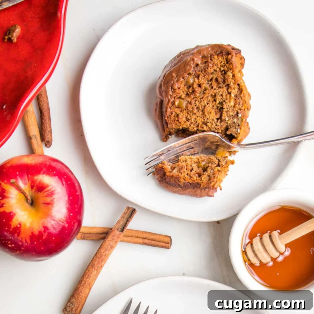 A perfectly portioned slice of moist apple honey cake, adorned with a luscious chocolate glaze, rests on a pristine white plate. In the background, a whole, ripe red apple, fragrant cinnamon sticks, and a small dish of golden honey complete this inviting tableau, highlighting the cake's key ingredients and deliciousness.