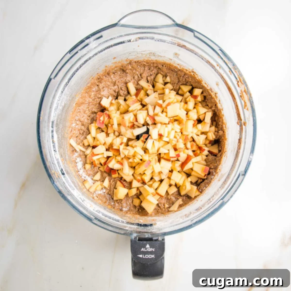 Thick cake batter nestled in the bowl of a food processor, with a generous layer of freshly chopped apple pieces carefully placed on top, ready to be folded in.