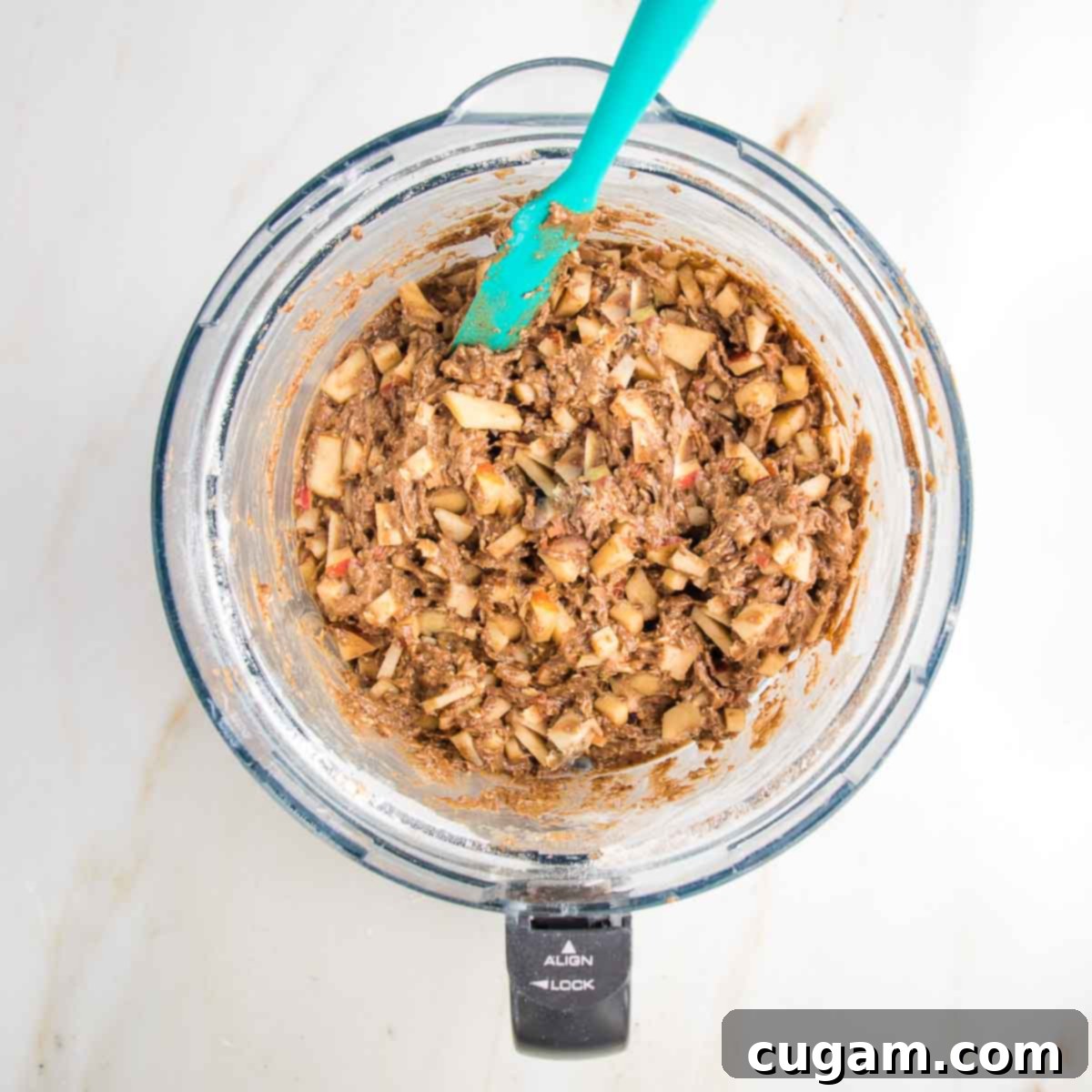 A rubber spatula gracefully mixing in chunks of diced apple into the thick, creamy cake batter within the food processor bowl, ensuring an even distribution of fruit throughout the cake.