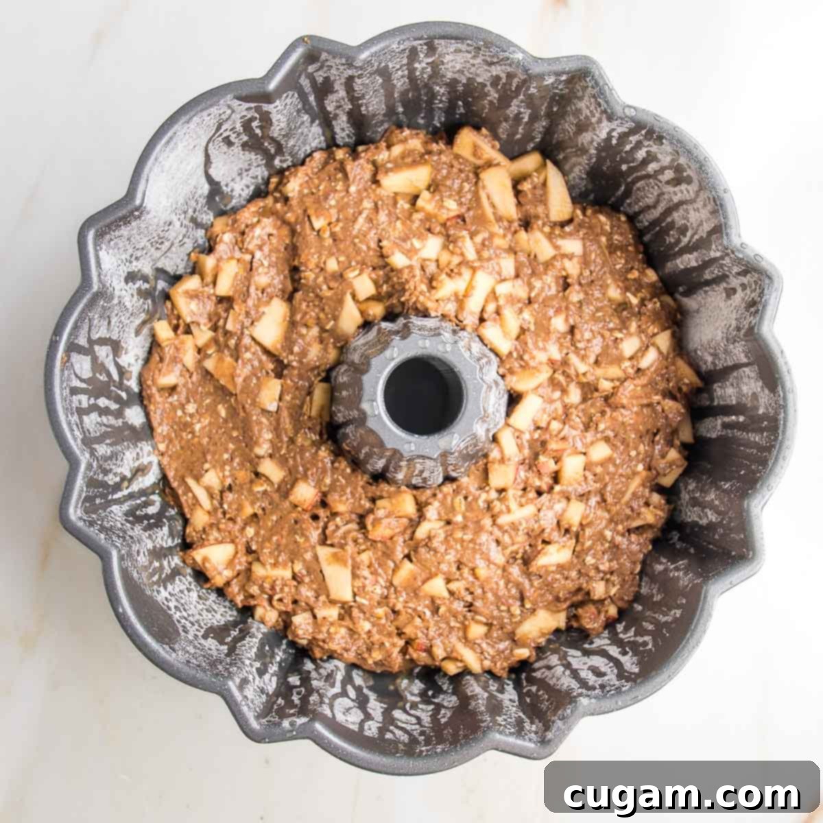 A bundt pan, meticulously prepared and filled with rich apple cake batter. The surface of the batter is dotted with visible chunks of diced apples, promising a delightful texture in every bite of the finished cake.