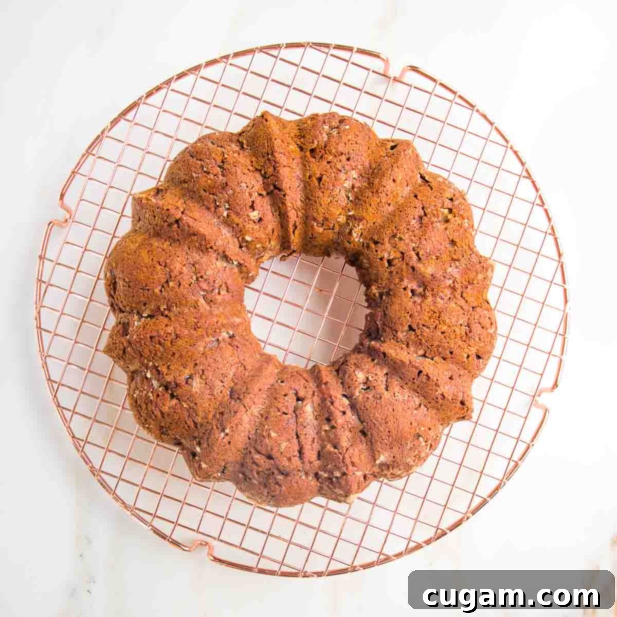 A golden-brown apple bundt cake, freshly baked, resting on a circular wire rack. The cake is perfectly shaped, indicating a successful release from the pan, and is cooling down to achieve its ideal texture before glazing.