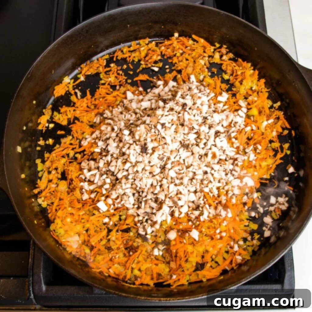 Cast iron skillet with sautéed veggies including shredded carrots. Small diced mushrooms are added to the top of the pan.