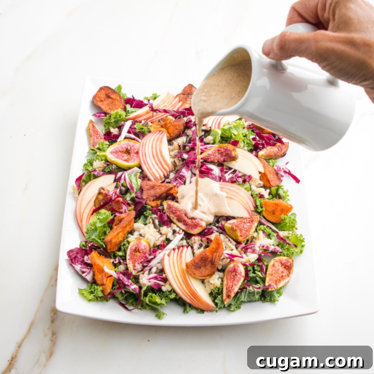 Creamy apple walnut dressing poured from white pitcher onto a platter filled with colorful salad. Greens, roasted sweet potatoes and figs, apple slices, wild rice.