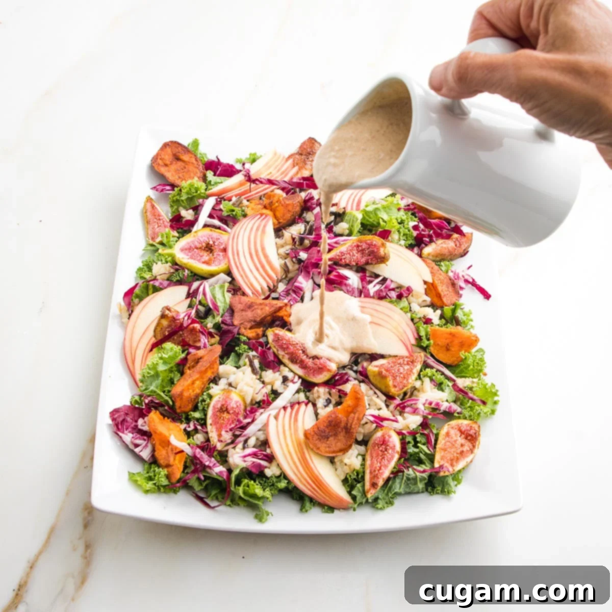 Creamy apple walnut dressing poured from white pitcher onto a platter filled with colorful salad. Greens, roasted sweet potatoes and figs, apple slices, wild rice.