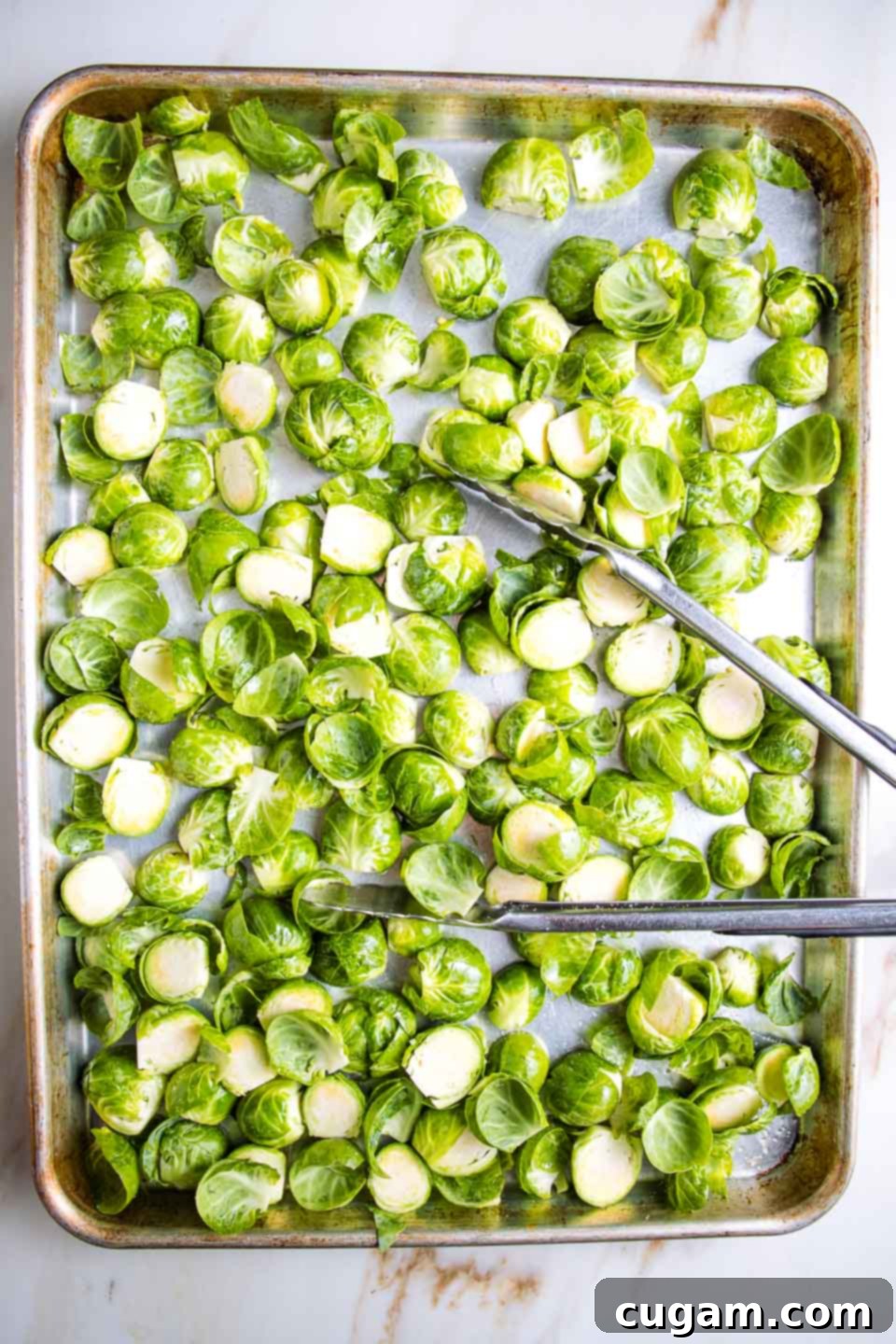 Large baking tray with tongs tossing halved Brussels sprouts.