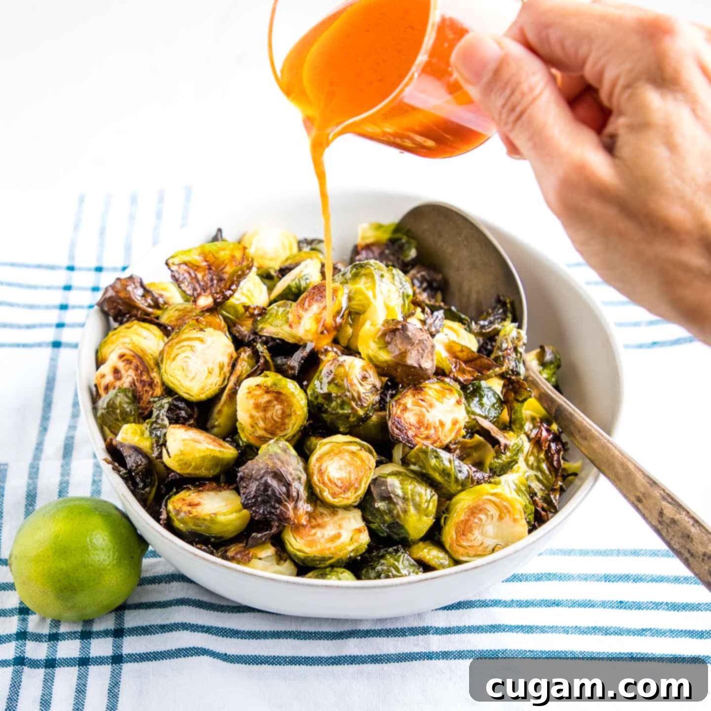 White bowl filled with roasted brussels sprouts and a pitcher of sweet and spicy sauce bring poured over and a lime on the side.