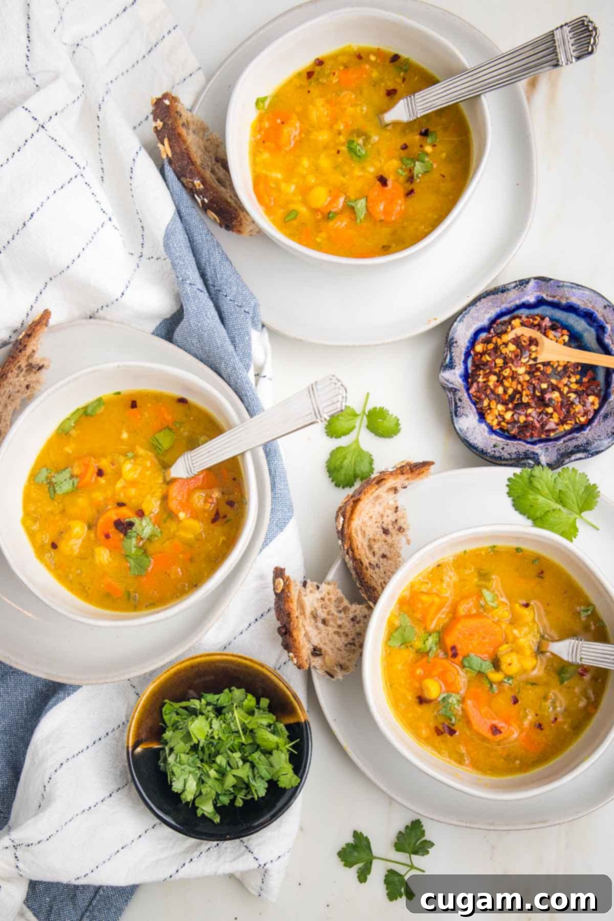 Bowls of yellow split pea soup, with crusty bread chunks, fresh parsley and crushed red pepper dispersed on a tea towel. 