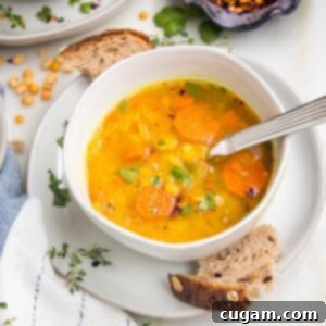 White bowl filled with yellow split pea soup with carrots and fresh herbs. Crusty bread pieces on the plate holding the soup bowl.