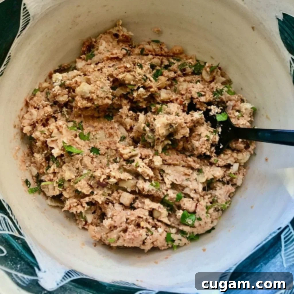 Beige bowl with a large black fork mashing together chickpeas, tomato paste, fresh parsley, and spices to form a flavorful vegan meatball mixture.