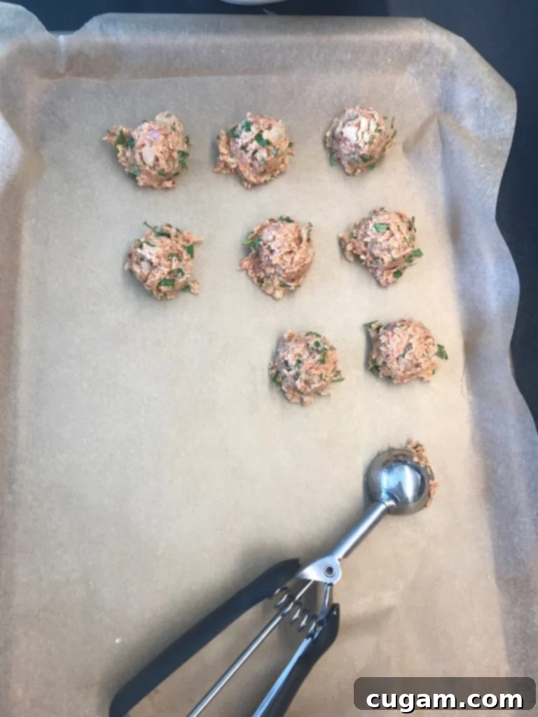 A large rimmed baking tray lined with unbleached parchment paper, with a small scoop meticulously portioning out vegan meatballs into neat, even rows.