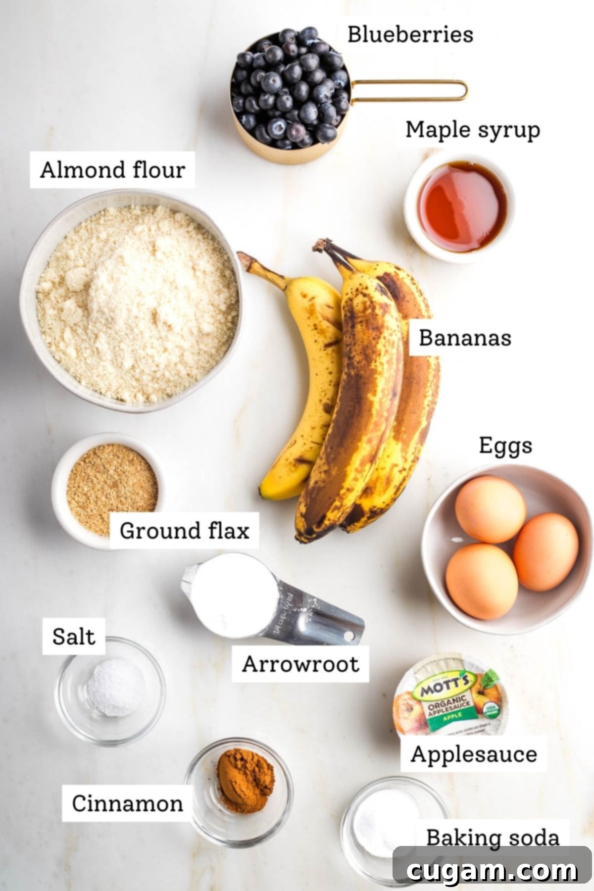 Individual ingredients for almond flour blueberry muffins laid out: fresh blueberries, almond flour, ripe bananas, eggs, ground flaxseed, cinnamon, arrowroot powder, unsweetened applesauce, and baking soda.