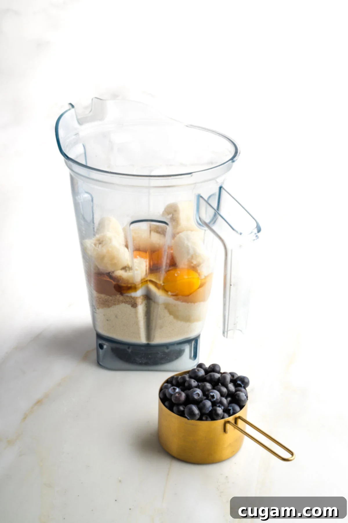 All the dry and wet ingredients for almond flour muffins, including bananas, almond flour, eggs, applesauce, arrowroot, flax, baking soda, cinnamon, and sea salt, gathered in a blender pitcher, with a separate cup of blueberries.