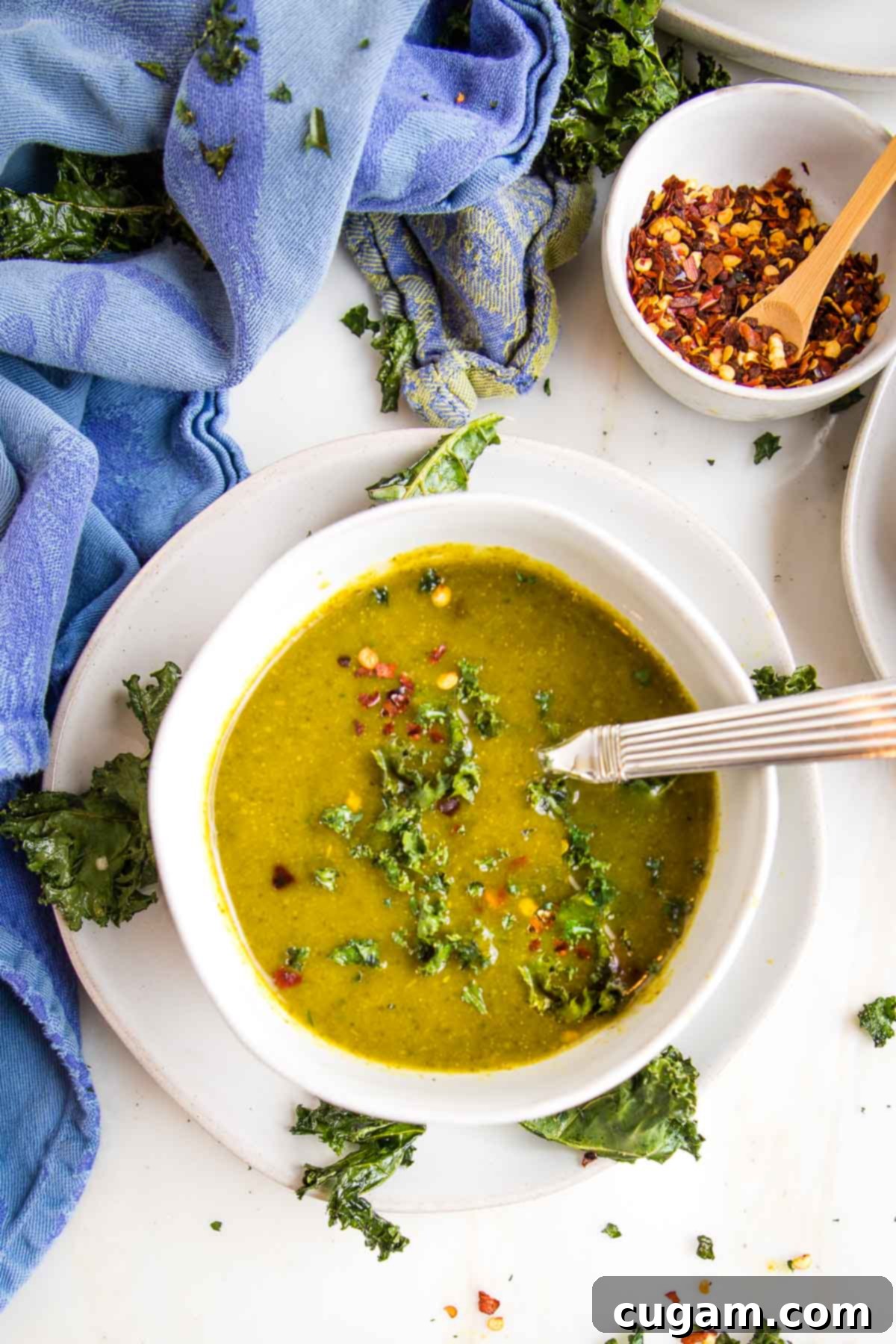 A rich, deep green creamy vegan parsnip and kale soup, elegantly served in a white bowl on a matching white saucer, garnished with crispy kale chips and a sprinkle of vibrant red pepper flakes.