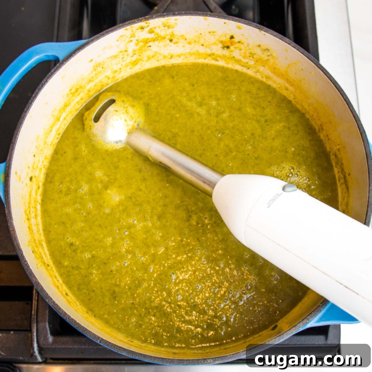 An immersion blender actively pureeing a vibrant green vegetable soup directly inside a blue Dutch oven, creating a smooth and creamy texture for the parsnip and kale soup.