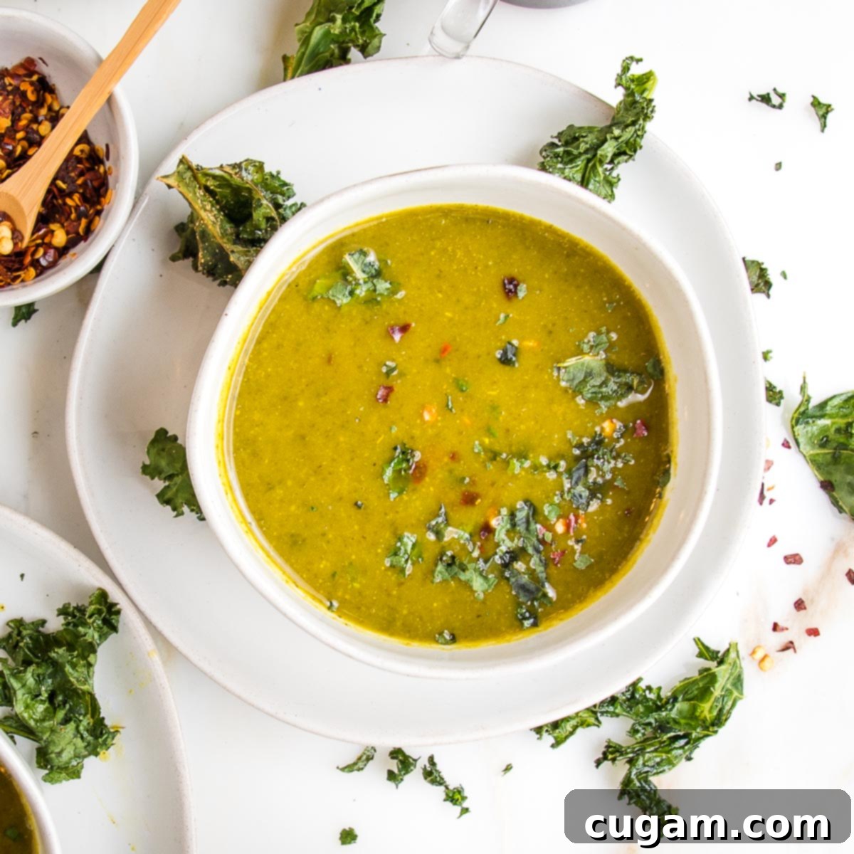 A pristine white bowl filled with a vibrant green pureed parsnip and kale soup, generously garnished with fresh herbs, crumbled crunchy kale chips, and a touch of red pepper flakes, creating an inviting and healthy meal.