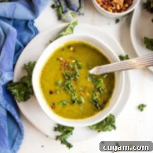 A white bowl filled with vibrant green pureed parsnip and kale soup, garnished with crispy kale chips and red pepper flakes, set on a white saucer with more kale chips.