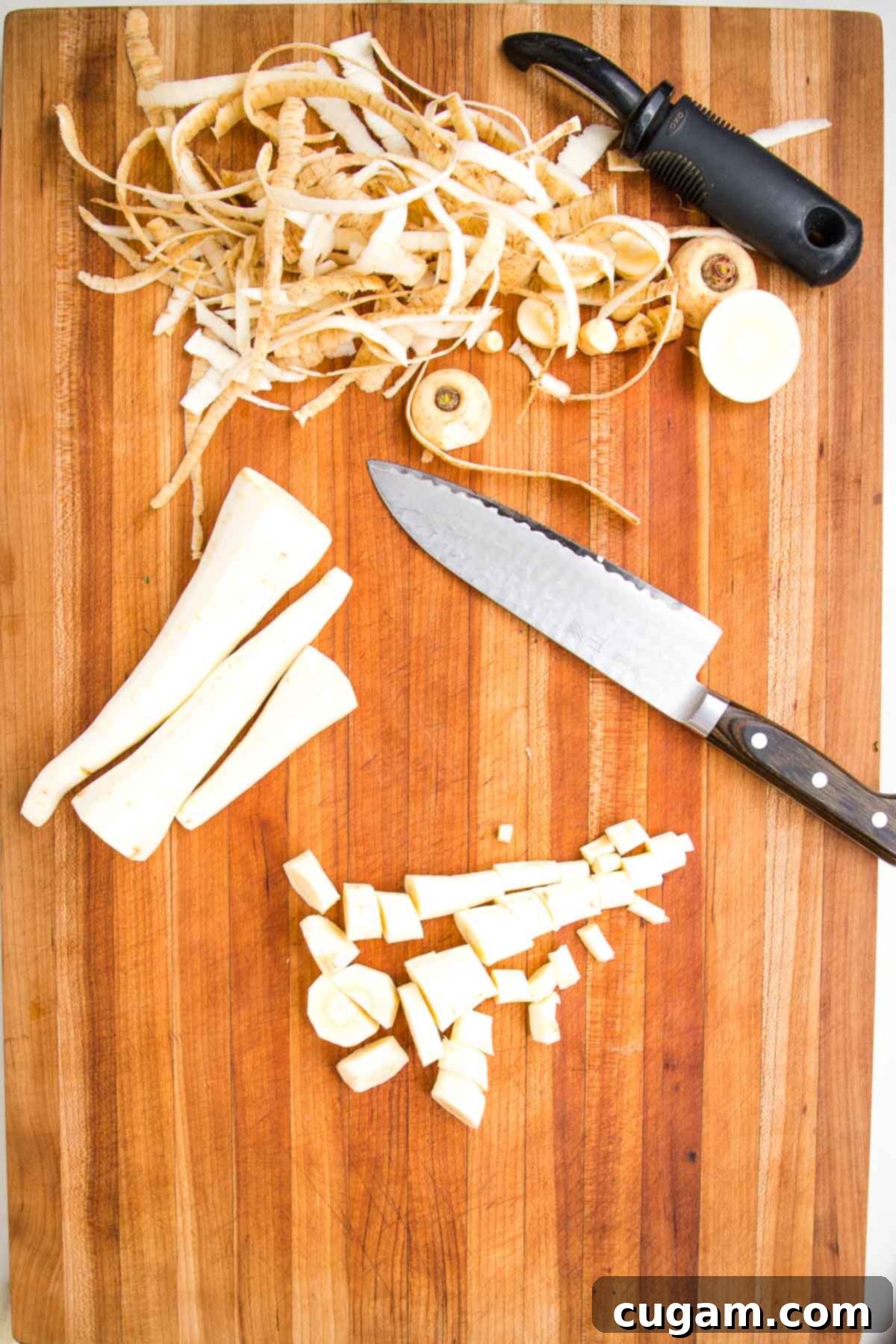 A wooden cutting board with a vegetable peeler, showcasing parsnip peels, three perfectly peeled parsnips, and a pile of roughly chopped parsnips alongside a chef's knife, illustrating the preparation process.