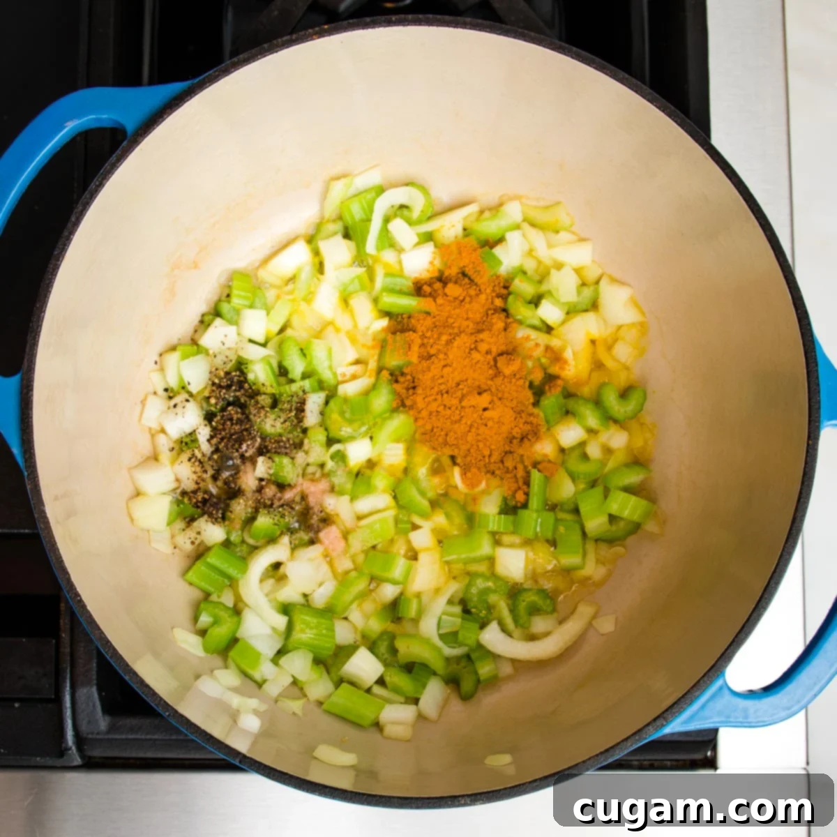 A Dutch oven filled with glistening, turmeric-infused sautéed onions and celery, seasoned with salt and pepper, with freshly chopped garlic resting on top, ready to be stirred in.